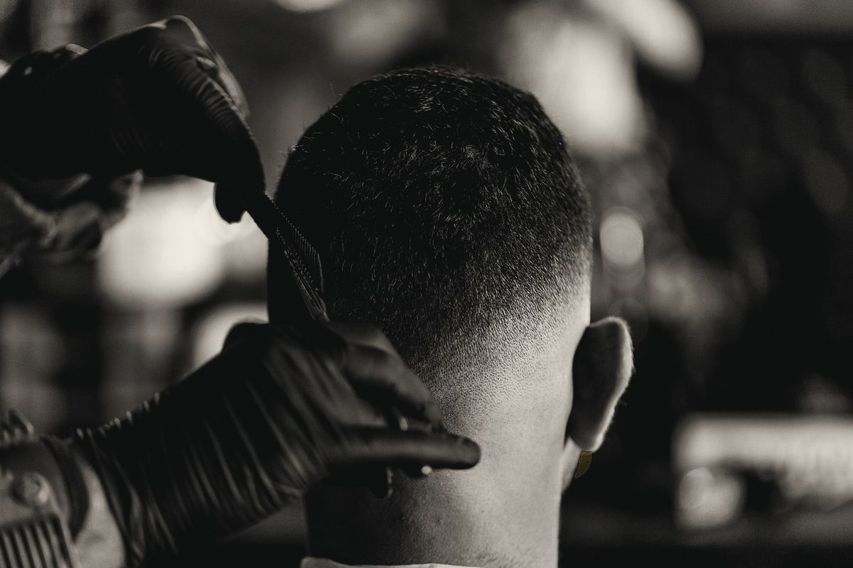A barber in black gloves finishing a skin fade at the back of the head with a comb and trimmer.
