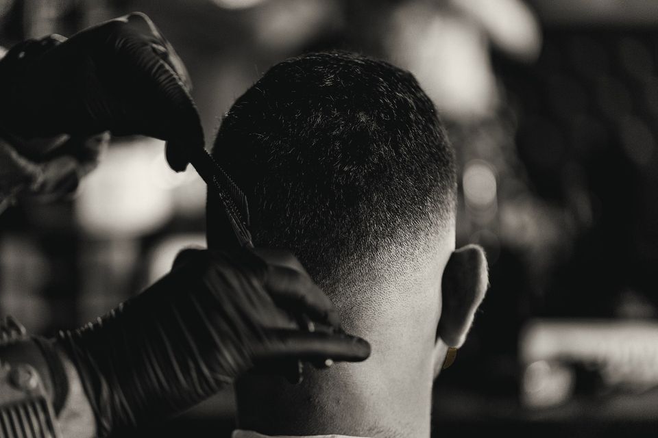 A barber in black gloves finishing a skin fade at the back of the head with a comb and trimmer.