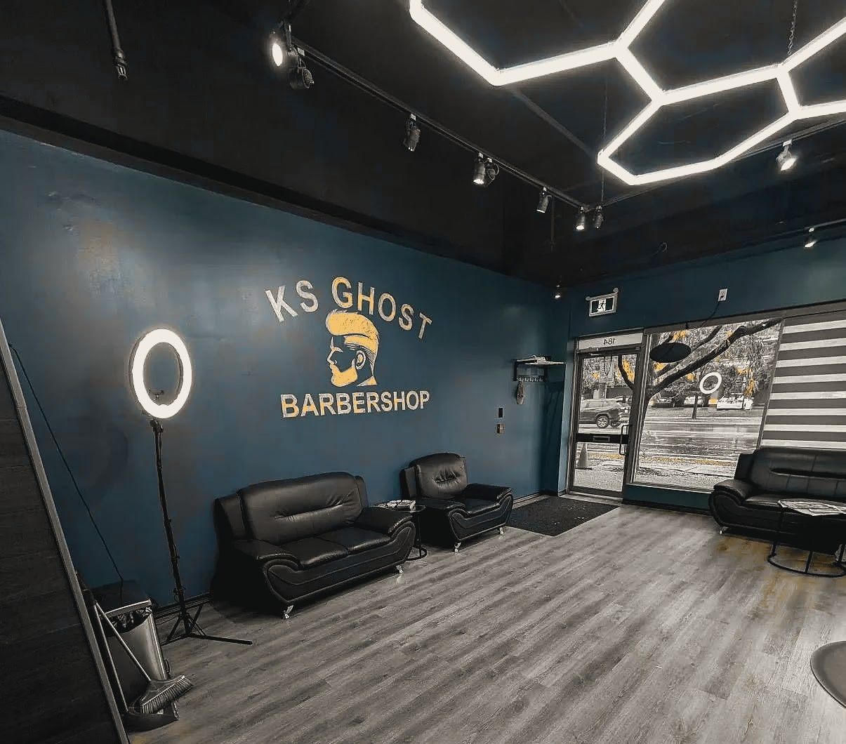 Inside KS Ghost Barbershop: the lounge with black leather sofas under the shop’s painted logo, hexagon lights overhead.