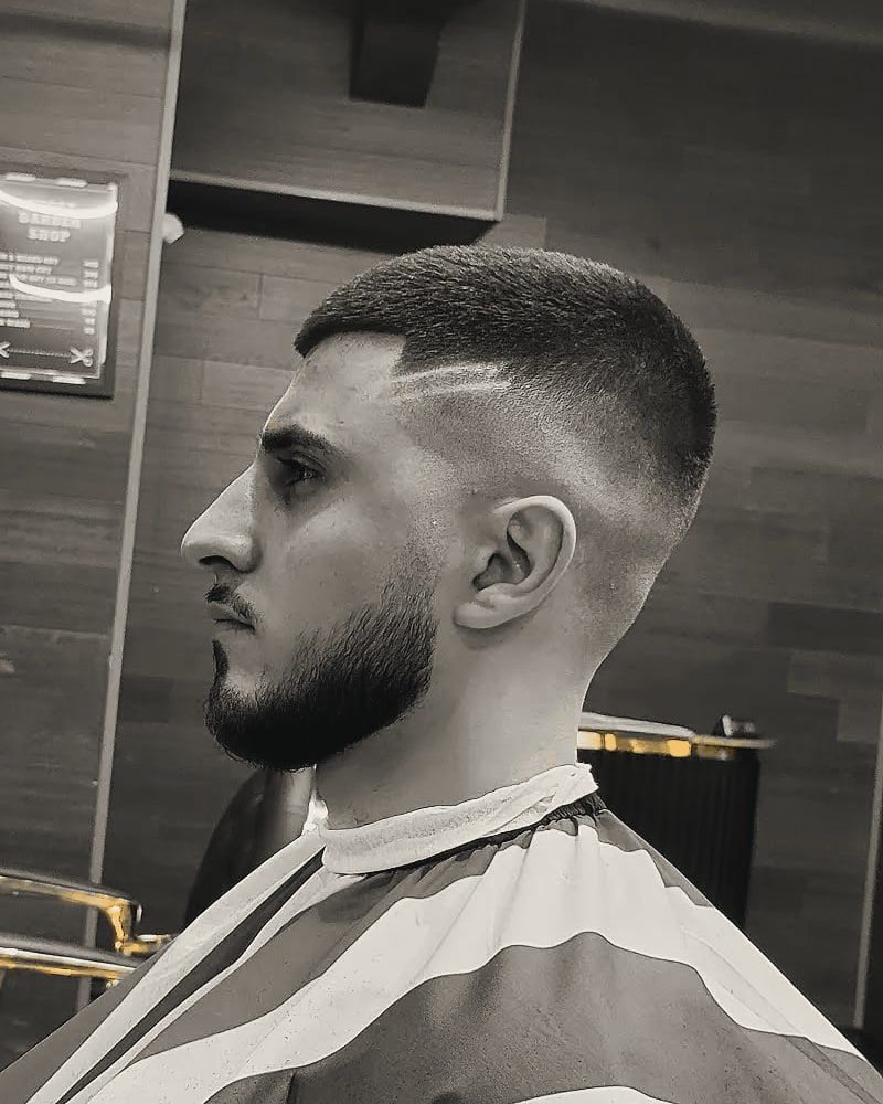 Profile of a short fade with a shaved part line and a full, sculpted beard.